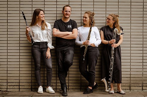 Vier Personen lehnen an einer Wand aus hellen Kacheln, drei halten Holzblasinstrumente, eine steht mit verschränkten Armen in schwarzer Kleidung.