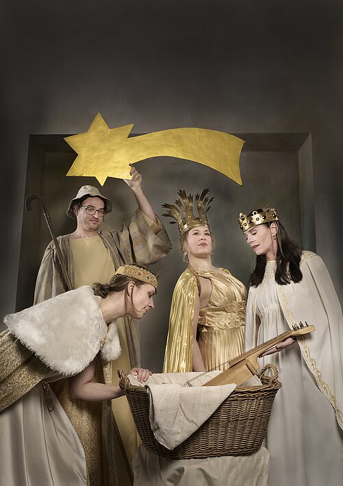 Drei Personen in historischen Gewändern stehen um eine mit Tuch bedeckte Wiege, darüber ein gelber Stern mit Schweif.