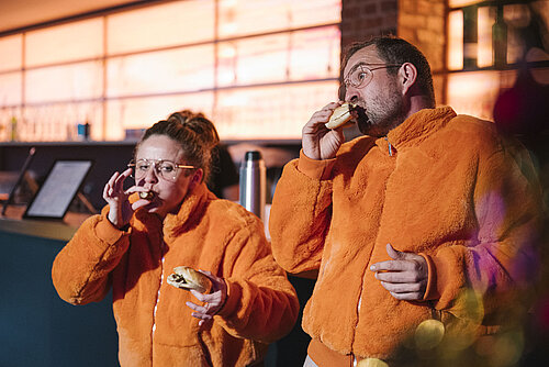 Zwei Personen in orangefarbenen Jacken essen und trinken in einem Innenraum mit warmem Licht.