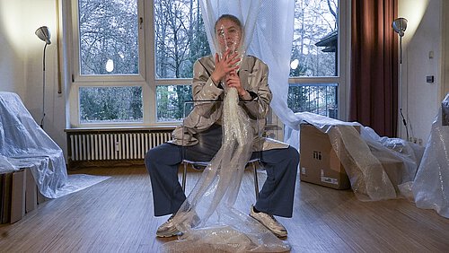 Eine Frau sitzt breitbeinig auf einem Stuhl und hält sich ein Luftpolster ans Gesicht 