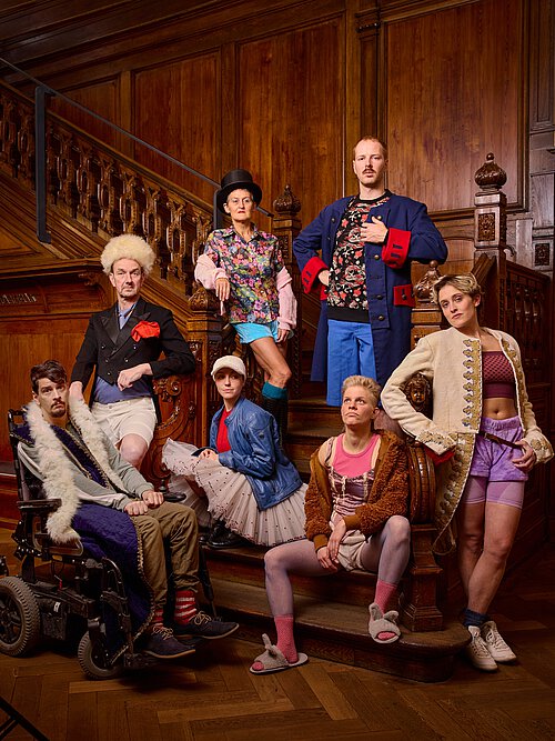 Gruppe in farbenfroher, ausgefallener Kleidung posiert auf einer Holztreppe mit geschnitztem Geländer in einem prunkvollen Raum.