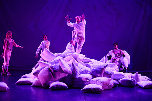 Mehrere Personen in Bühnenkostümen um einen großen Haufen Kissen, eine Person springt von oben herunter, violettes Bühnenlicht.