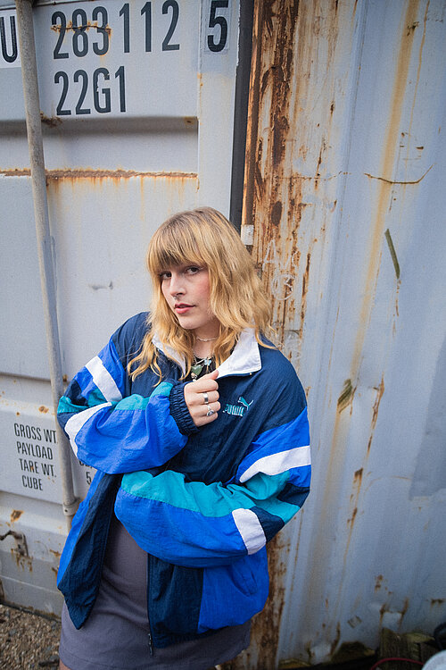 eine junge Frau mit Trainingsjacke vor einem grauen Container