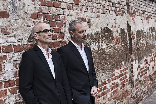 Zwei Männer in schwarzen Anzügen lehnen an einer alten Backsteinmauer mit Putzstellen.