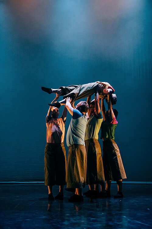 Gruppe von fünf Tänzern hebt einen weiteren Tänzer waagerecht über ihren Köpfen auf einer Bühne mit blauem Hintergrund