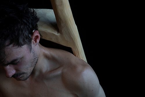 Oberkörper eines nackten Mannes mit dunklen, lockigen Haaren und silbernem Ohrstecker vor schwarzem Hintergrund.