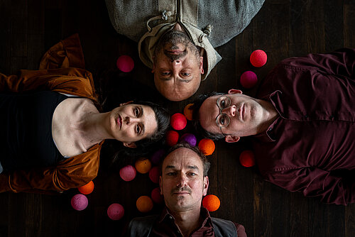Drei Männer und eine Frau liegen auf einem dunkeln Holzboden. Um sie herum liegen bunte Bälle verstreut.