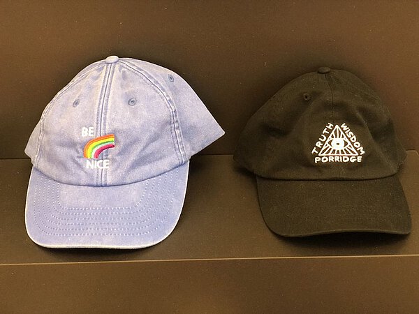 Baseball-Caps von David Shrigley in Blau mit Regenbogen (Be Nice) und Schwarz mit einem Dreieck mit Auge. An den Seiten beschriftet mit Thruth, Wisdom, Porridge