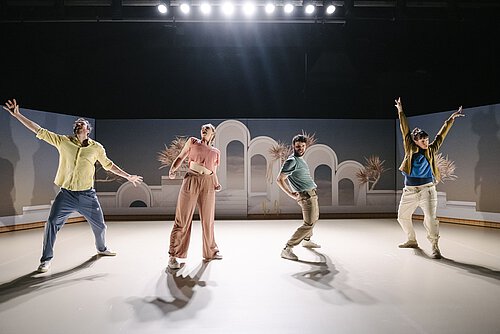An der Bühnenkante zwei Männer und zwei Frauen in energetischen Posen, im Hintergrund Gegenlicht der Scheinwerfer, auf der Bühnenrückwand pastellfarbiges Fotomotiv (antike Bögen und Palmen)