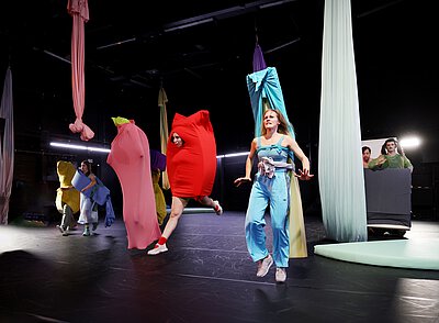 Mehrere Personen in bunten, teils figurbetonten Kostümen bewegen sich auf einer Bühne mit hängenden Stoffbahnen.