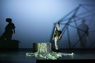 Person mit einer militärischen Weste, die über die Bühne geht.   Im Hintergrund eine weiße Leinwand, auf die Schatten der Person und einer geometrischen Form geworfen werden. Die Beleuchtung ist hell und diffus, mit einem bläulichen Farbton. Die Person schaut fokussiert in eine Richtung. Die Atmosphäre ist düster und ernst.