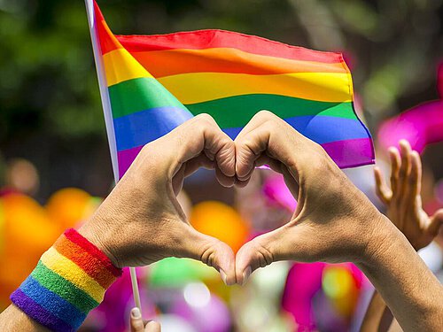 zwei Händer, deren Finger ein Herz formen vor Regenbogenflagge