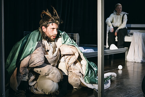 Mann in Käferkostüm in metallener Binnenkadrierung sitzend, im Hintergrund eine Frau in Anzug sitzend 