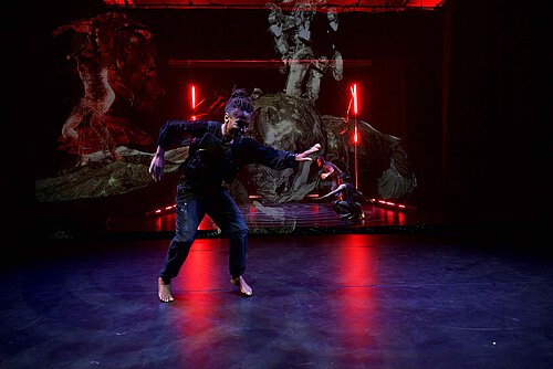 Tanzender Performer in schwarzem Oberteil und Jeans auf einer dunklen Bühne mit roten Lichtern und abstrakten Projektionen im Hintergrund.