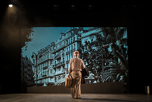 ein Mann mit Koffer in der Hand und Jackett über seinem Arm schaut auf Gebäude, die an die Wand projiziert werden
