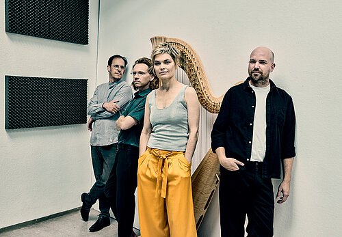 Vier Personen, im Vordergrund eine Frau mit kurzen blonden Haaren, orangfarbener Hose und grauem Trägertop. Dahinter steht eine große Konzertharfe