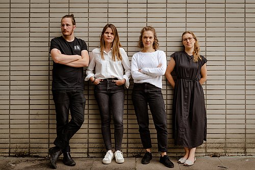 Vier Personen lehnen an einer Wand aus hellen Kacheln, zwei Frauen tragen schwarze Hosen, eine Frau ein schwarzes Kleid, ein Mann schwarze Kleidung.