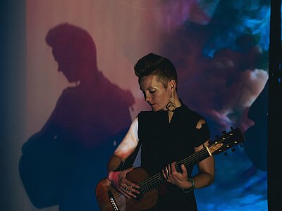 Person mit kurz geschnittenem Haar spielt akustische Gitarre vor einer Wand mit buntem Licht und Schatten.