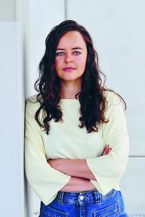 Person mit langen lockigen Haaren lehnt mit verschränkten Armen an einer Wand und trägt ein helles Oberteil mit weiten Ärmeln und blaue Jeans