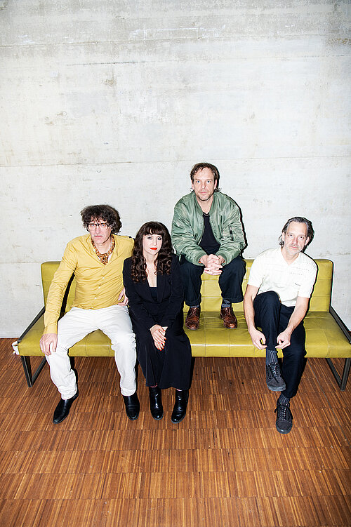 Vier Personen sitzen und hocken auf einem grünen Sofa vor einer Betonwand, alle tragen unterschiedliche Outfits und blicken zur Kamera.