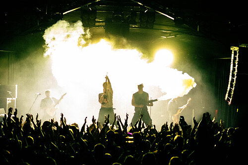 Band spielt auf Bühne vor großem Publikum mit hellen Bühnenlichtern und Rauch