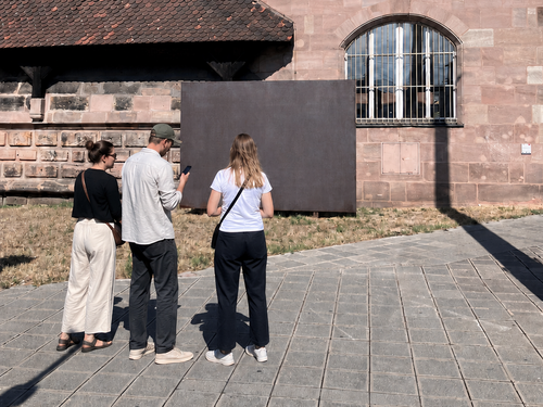 Drei Personen stehen vor einem hohen, pinken Fahnenmast mit wehender pinker Fahne neben einem historischen Sandsteingebäude im öffentlichen Raum