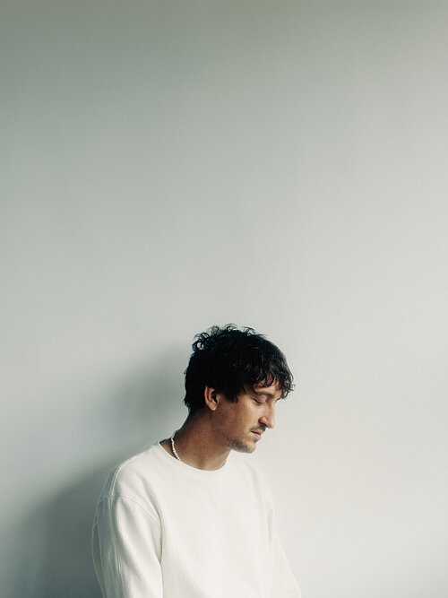 ein Mann mit weißem Pulli vor weißem Hintergrund, der Kopf ist gesenkt