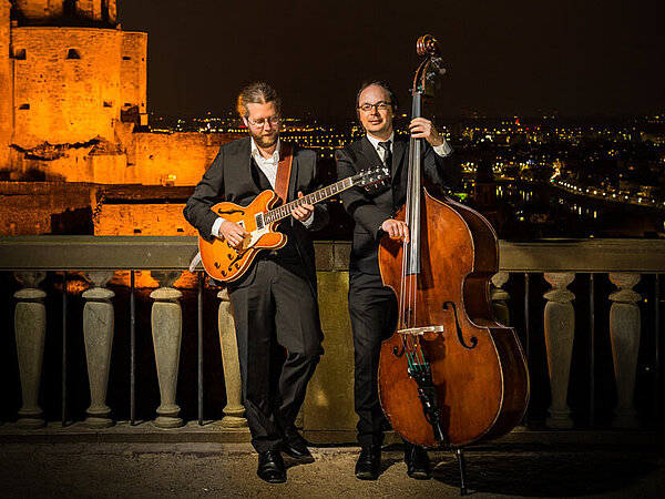 Die beiden Musiker von Gramm Art Project stehen vor einer nächtlichen Stadtansicht