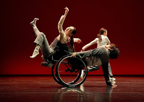 Drei Tänzer auf Bühne vor rotem Hintergrund, einer davon sitzt im Rollstuhl und hebt die Arme, die anderen beiden bewegen sich dynamisch.