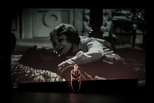 Ein Mann sitzt vor einer Leinwand, auf der eine Projektion in schwarz-weiß mit einer Frau zu sehen ist
