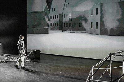 Person mit Rucksack steht auf einer Bühne vor einer großflächigen Projektion von Häusern