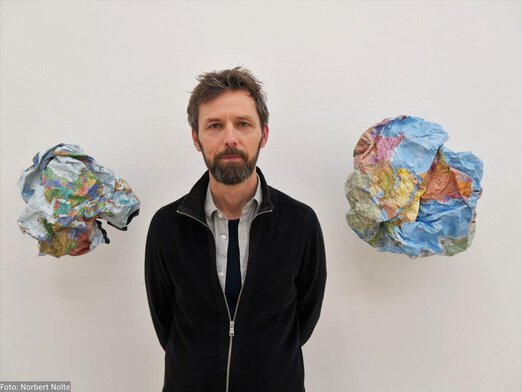 Oliver van den Berg steht zwischen zwei seiner Wandobjekten Knitterwelt 2 und 4. Das sind zwei kugelförmig zerknüllte Weltkarten aus Papier