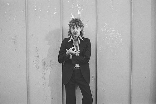 Auf dem schwarz-weiß Bild ist ein Mann in Anzug zu sehen, der in die Kamera blickt und vor seiner Brust mit Daumen und Zeigefinger einen Kreis bildet. 