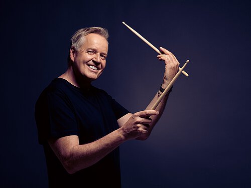 lachender Mann mit schwarzem T-Shirt und Drumsticks in der Hand
