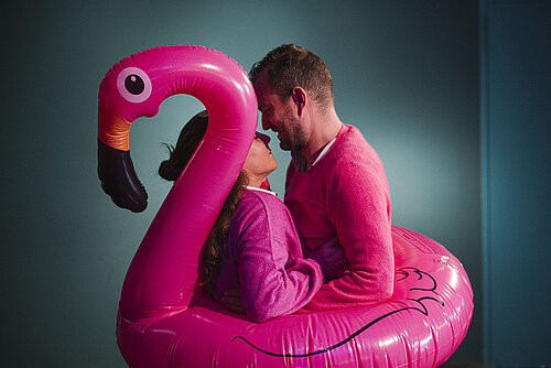Zwei Personen in pinker Kleidung sitzen in einem aufblasbaren pinken Flamingo mit schwarzem Schnabel.