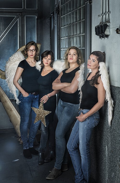 Vier Personen in schwarzer Oberbekleidung und blauer Jeans stehen in einem industriellen Raum mit grauen Wänden und Metallgittern.