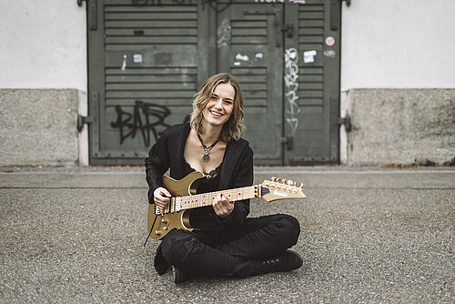Eine Frau in schwarzer Kleidung sitzt auf dem Boden und spielt E-Gitarre, sie lächelt dabei