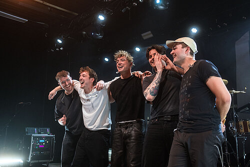 Fünf Männer stehen eng nebeneinander auf einer Bühne mit Bühnenbeleuchtung im Hintergrund, einige tragen T-Shirts und eine Person eine Kappe