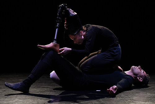 Zwei Tänzer in schwarzer Kleidung auf dunklem Bühnenboden, einer liegt auf dem Rücken und hält das Bein des anderen mit einer Prothese hoch
