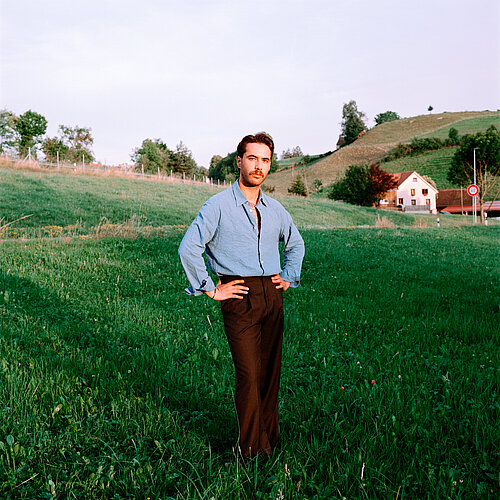 Ein Mann steht auf einer Wiese, im Hintergrund ein Hügel und Häuser