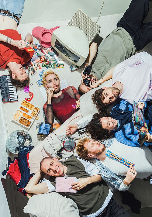 Sieben Personen liegen umringt von Essen und verschiedenen elektronischen Geräten auf dem Boden. Die Kamera fotografiert von oben.