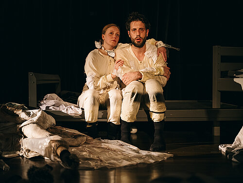 Mann und Frau beide in weißem Overall Arm in Arm sitzend auf metallenem Bettgestell