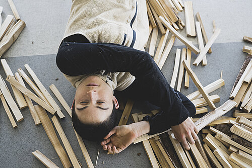 Junge Person liegt auf dem Rücken auf dem Boden, umgeben von verstreuten Holzstäben, trägt helles Oberteil mit dunklen Ärmeln.