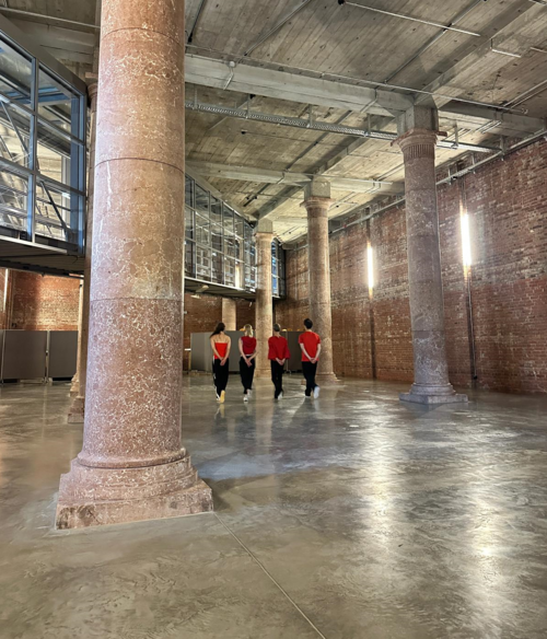 Vier Personen in roter Oberbekleidung gehen in einem großen Raum mit hohen Säulen und Backsteinwänden weg vom Betrachter