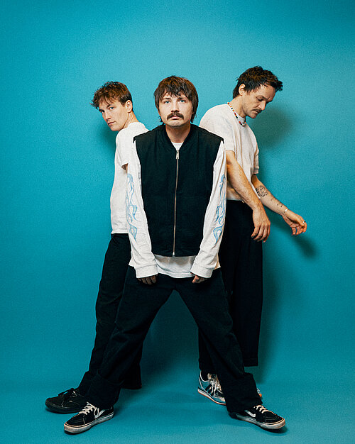 Drei Männer in lässiger Kleidung stehen vor einem türkisem Hintergrund