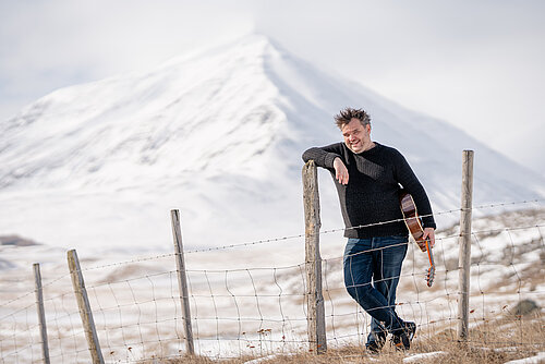 Mann in dunkler Kleidung lehnt an Zaunpfahl mit Gitarre unter dem Arm