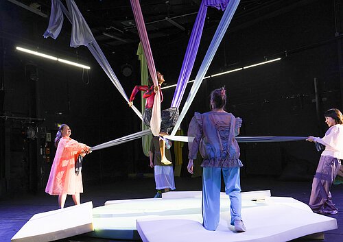 Person sitzt in einem pinken Oberteil auf einem weißen Tuch, das von mehreren Personen gehalten wird, auf einer weißen Bühne in einem dunklen Raum