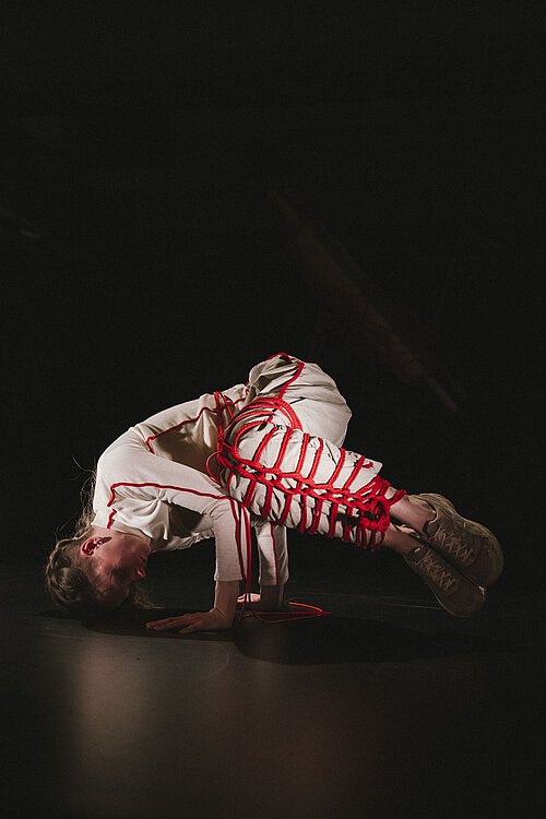 Eine Frau macht einen niedrigen Handstand, ihre beiden sind mit rotem Garn umhäkelt