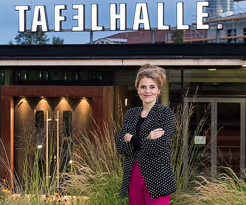 Person mit hochgestecktem Haar, gepunktetem Blazer und pinker Hose steht mit verschränkten Armen vor einem Gebäude mit dem Schriftzug 'TAFELHALLE'