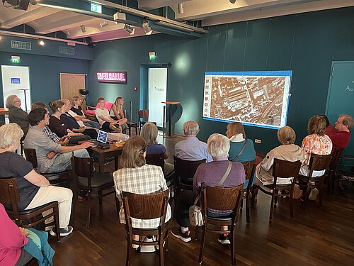 Gruppe von Menschen sitzt auf Stühlen in einem Raum und schaut auf eine Wandprojektion mit einer Luftaufnahme.